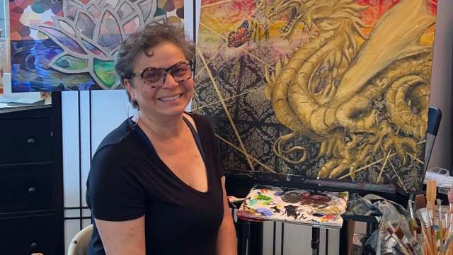 Artist Jill Covell, with paint brush in hand, sits in front of two of her paintings, including one showing a butterfly and a dragon.