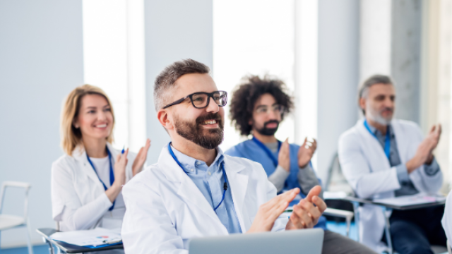 The image shows a diverse group of healthcare professionals, possibly at a meeting, applauding.