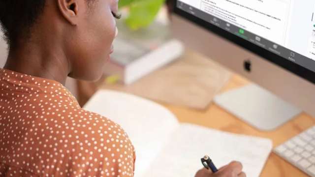 woman writing on notebook at desk with a computer