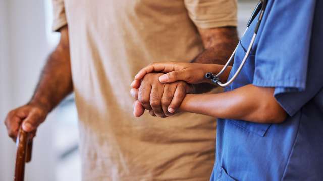 A caregiver holds a patient's hand, helping the patient to walk.