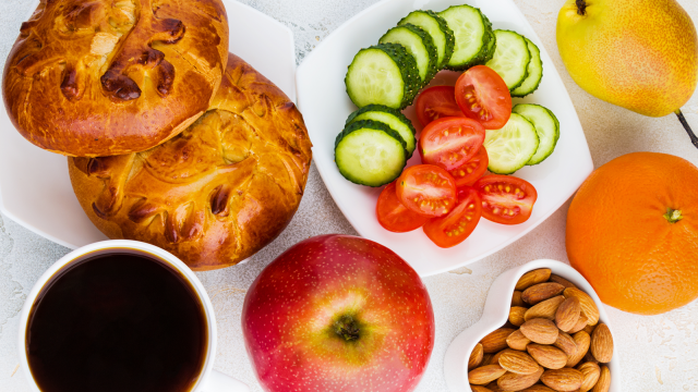 Balanced meal with bread rolls, sliced cucumber and tomato, almonds, fruit, and coffee, promoting healthy nutrition habits.