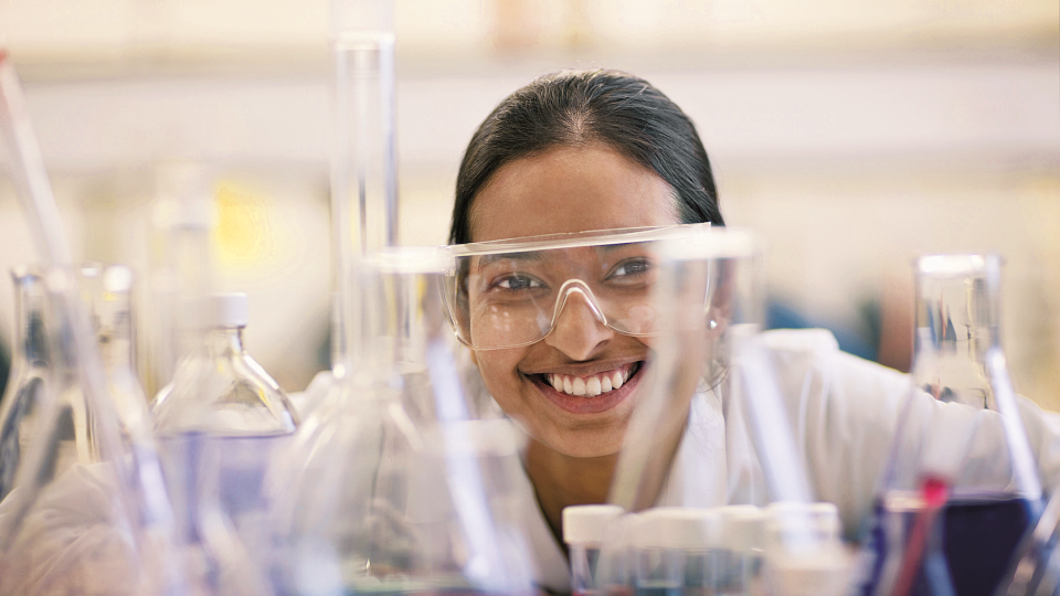 Woman smiling in lab wearing protective glasses surrounded by beakers and chemicals.