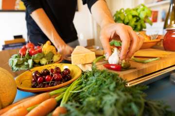 A person stands at a table, preparing a meal with an assortment of colorful vegetables and fruits laid out.