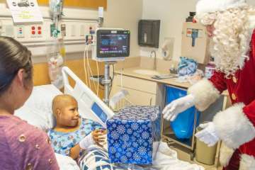 Arturo in a hospital bed receiving a gift from Santa while a woman watches.