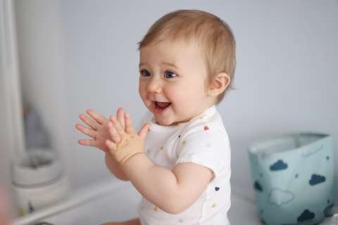 Baby in white onesie clapping hands beside a mirror and blue storage bin