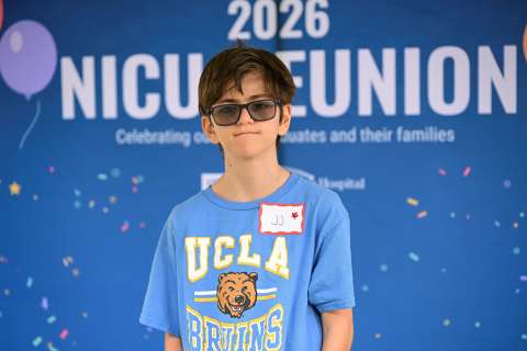 A boy wearing a UCLA T-shirt stands in front of a 2026 NICU Reunion sign.
