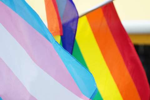A close-up of the Transgender Pride Flag (blue, pink, white) and the Rainbow Pride Flag (red, orange, yellow, green, blue, purple) flying together.