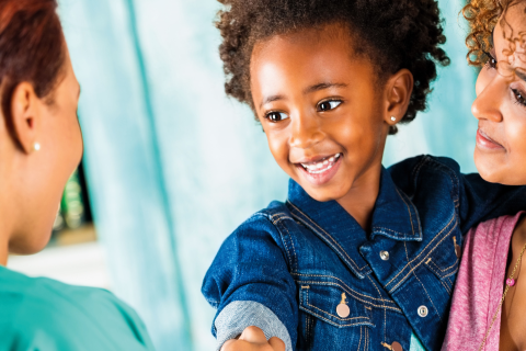 A smiling young child in a denim jacket, held by a parent, shakes hands with a healthcare provider.