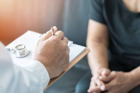 Person in white coat writing on clipboard as seated patient waits