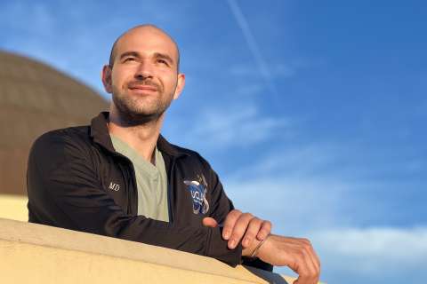 A doctor leans on a wall looking out into the distance, with the blue sky above.