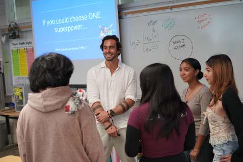 A man smiles while talking with a small group of students in a classroom, standing in front of a whiteboard and a projected slide that reads "If you could choose ONE superpower... What would you choose? Why?"