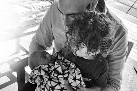 Black and white photo: An adult man holds a swaddled newborn baby while being embraced by a young child with curly hair in a bright, outdoor setting.