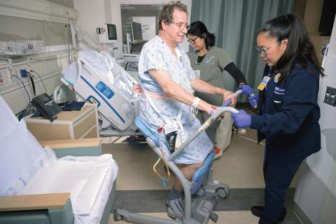 Two nurses assist a patient in a hospital room using a patient lift.