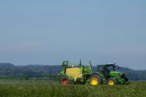 Green tractor spraying crops in a field under a clear blue sky.