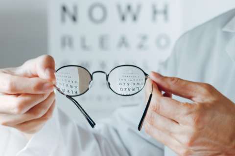 Person in white coat holding round eyeglasses in front of blurred eye chart