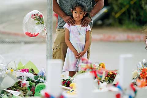A young girl stands in front of a memorial covered in flowers, with an adult behind her.
