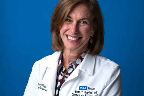 Dr. Beth Karlan smiling and wearing her UCLA lab coat with a blue backdrop behind.