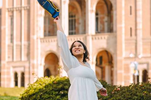 Neha Pai smiling, tossing graduation cap in front of a university building