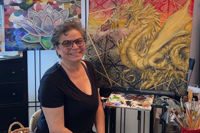 Artist Jill Covell, with paint brush in hand, sits in front of two of her paintings, including one showing a butterfly and a dragon.