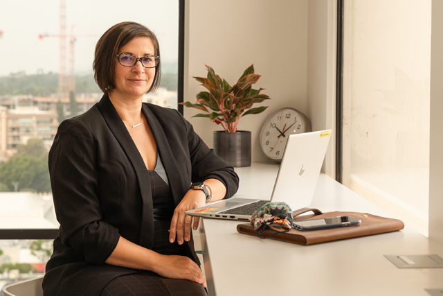 Director of Cancer Genetic Counseling, Mariana Niell-Swille, posing on her office wearing a black dress with a black jacket on top, and her laptop in front of her