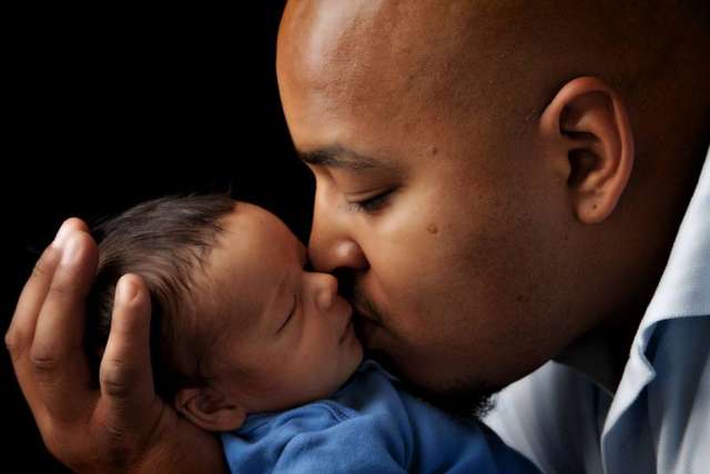Man cradles a baby's head against a dark background.