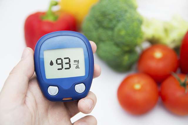 Hand holding a blue glucose meter displaying 93 mg/dL, with fresh vegetables like tomatoes, broccoli, and bell peppers in the background.