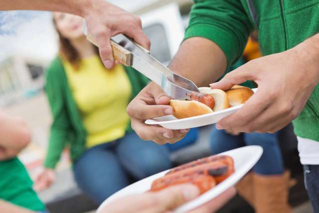 A person in a green shirt uses tongs to place a grilled hot dog onto a bun held by another person during an outdoor gathering or tailgate.