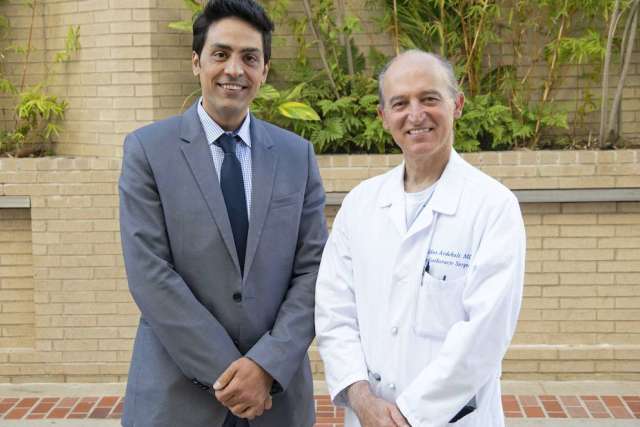 Two men smile while standing outdoors against a brick wall and plants: one in a gray suit and tie, and the other in a white lab coat.