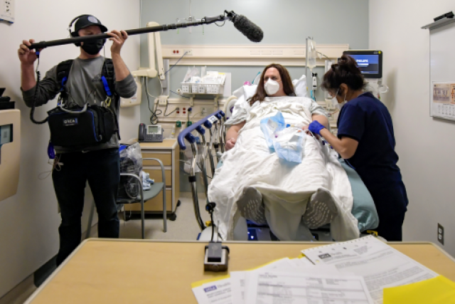 A cameraperson records Chloe while masked and lying in a hospital bed while a nurse tends to her.