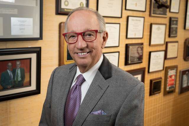 Ronald W. Busuttil, MD, PhD standing in front of a yellow wall with many picture frames, smiling and looking at the camera
