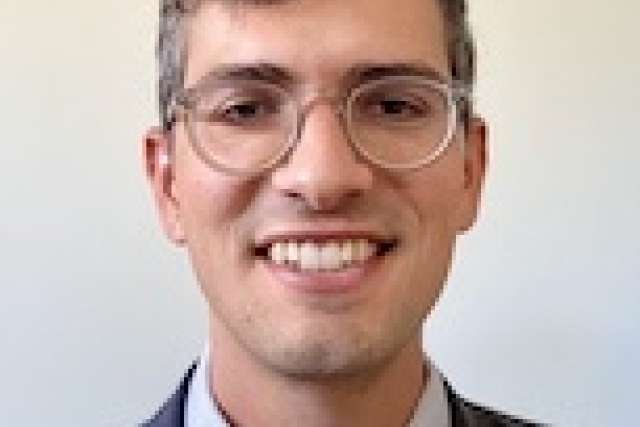  A headshot of Daniel Stokes, a smiling man with short curly hair and glasses, wearing a suit and patterned tie.