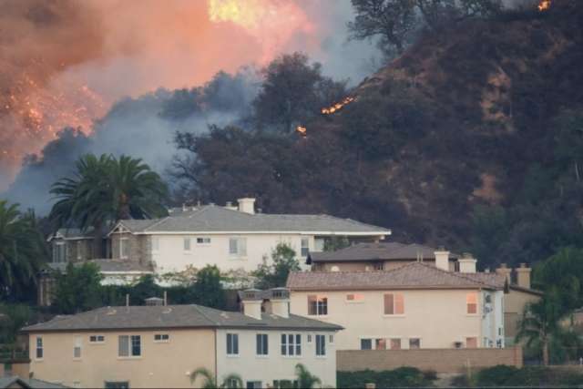 Wildfire burning on a hillside near residential homes, with flames and smoke visible among trees.