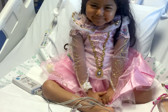 Young girl in a pink princess dress smiling while sitting cross-legged in a hospital bed at UCLA Health.