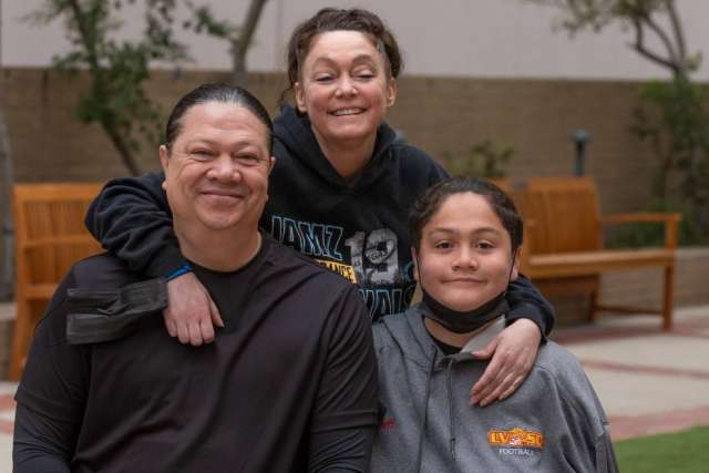 Three people wearing casual hoodies sitting and standing together outdoors near benches and greenery.