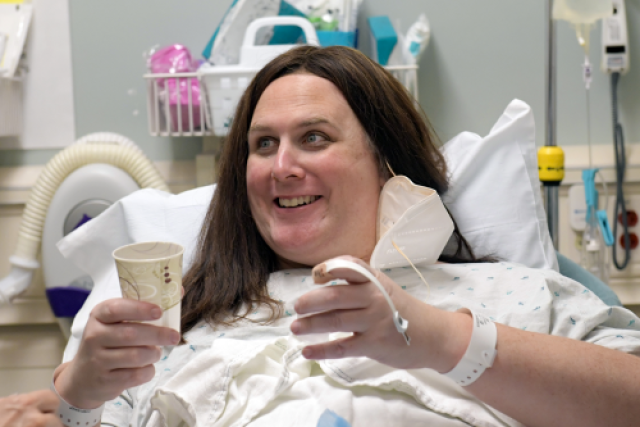 Chloe smiling with long brown hair, wearing a hospital gown and a white wristband, sits up in bed with a face mask hanging from one ear.