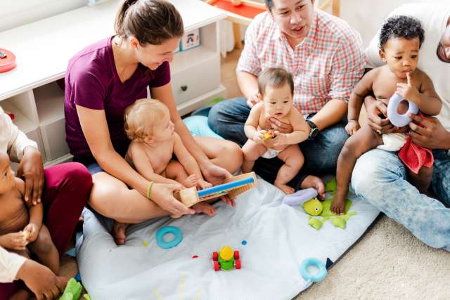 Babies and adults seated in a circle on a blanket sharing toys.
