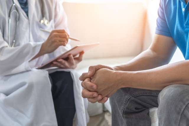 A doctor taking notes while a patient sits with clasped hands.