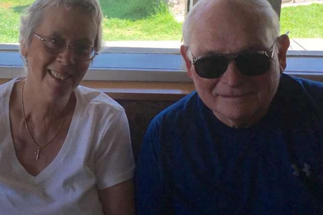 Two people sitting indoors near a window with sunlight coming through, one wearing a white shirt with a necklace and the other in a dark shirt.