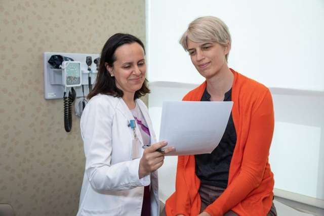 Lori Jordan, in an orange cardigan, reviews a paper with a woman doctor wearing a white coat in an exam room.