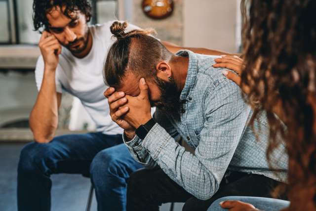 A man is consoled by two other members of a support group, all sitting in a circle.