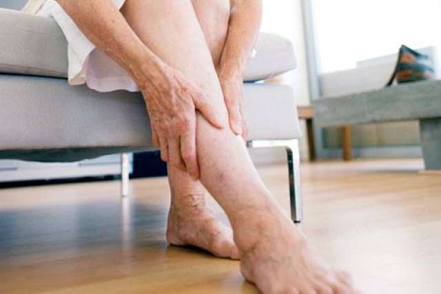 Person sitting on a chair, holding their ankle with one hand, bare feet resting on a wooden floor.