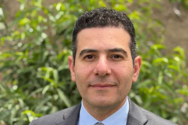 Professional headshot: man in suit and tie, green foliage background.
