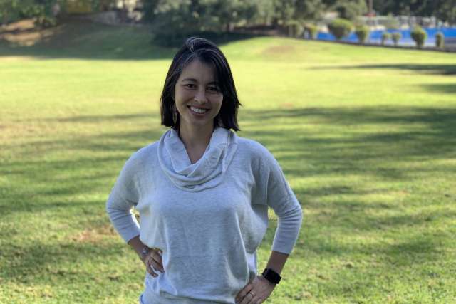 A headshot of Camila Guerrero, a speech therapist with dark hair and a light sweater, standing and smiling confidently outdoors on a grassy field.