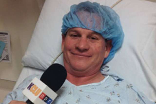 Lance Orozco in hospital gown and surgical cap lying in bed speaking to a microphone