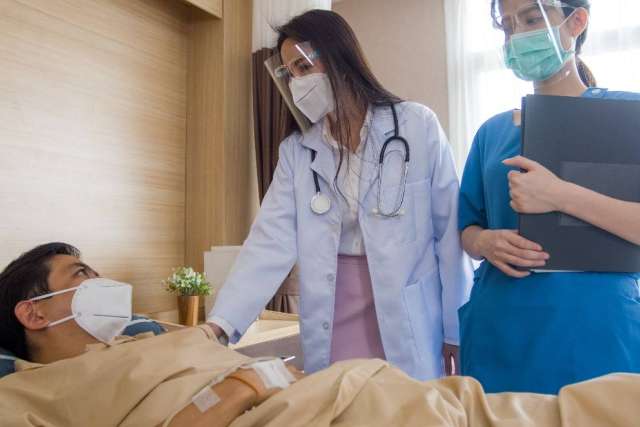 Two healthcare professionals standing by a hospital bed, one with a stethoscope and the other holding a folder, attending to a patient.