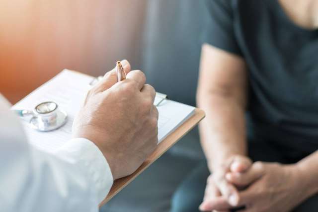 Person in white coat writing on clipboard as seated patient waits