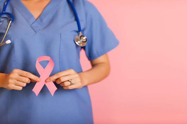 A healthcare worker in blue scrubs and a stethoscope holds a pink awareness ribbon in front of their chest against a solid pink background.