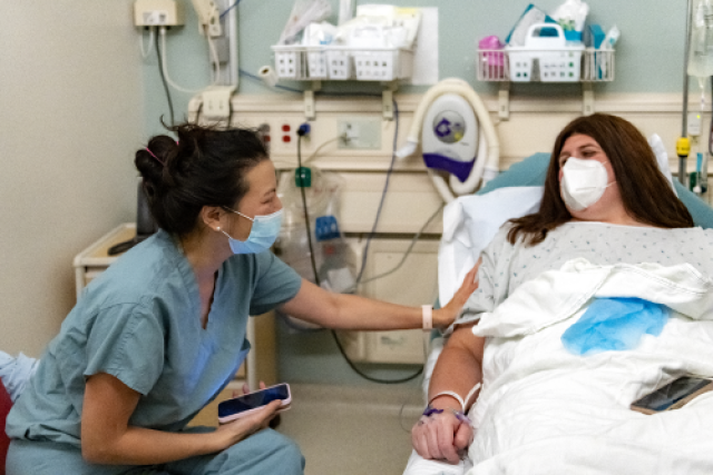 A masked nurse in teal scrubs comforts a masked patient named Chloe in a hospital gown lying in a bed.