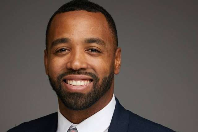 Person wearing a dark suit jacket, white dress shirt, and a patterned tie against a plain gray background.