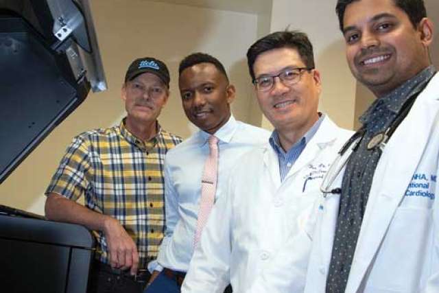 Group of people standing together indoors, including individuals wearing white medical coats and stethoscopes, next to a large medical device.